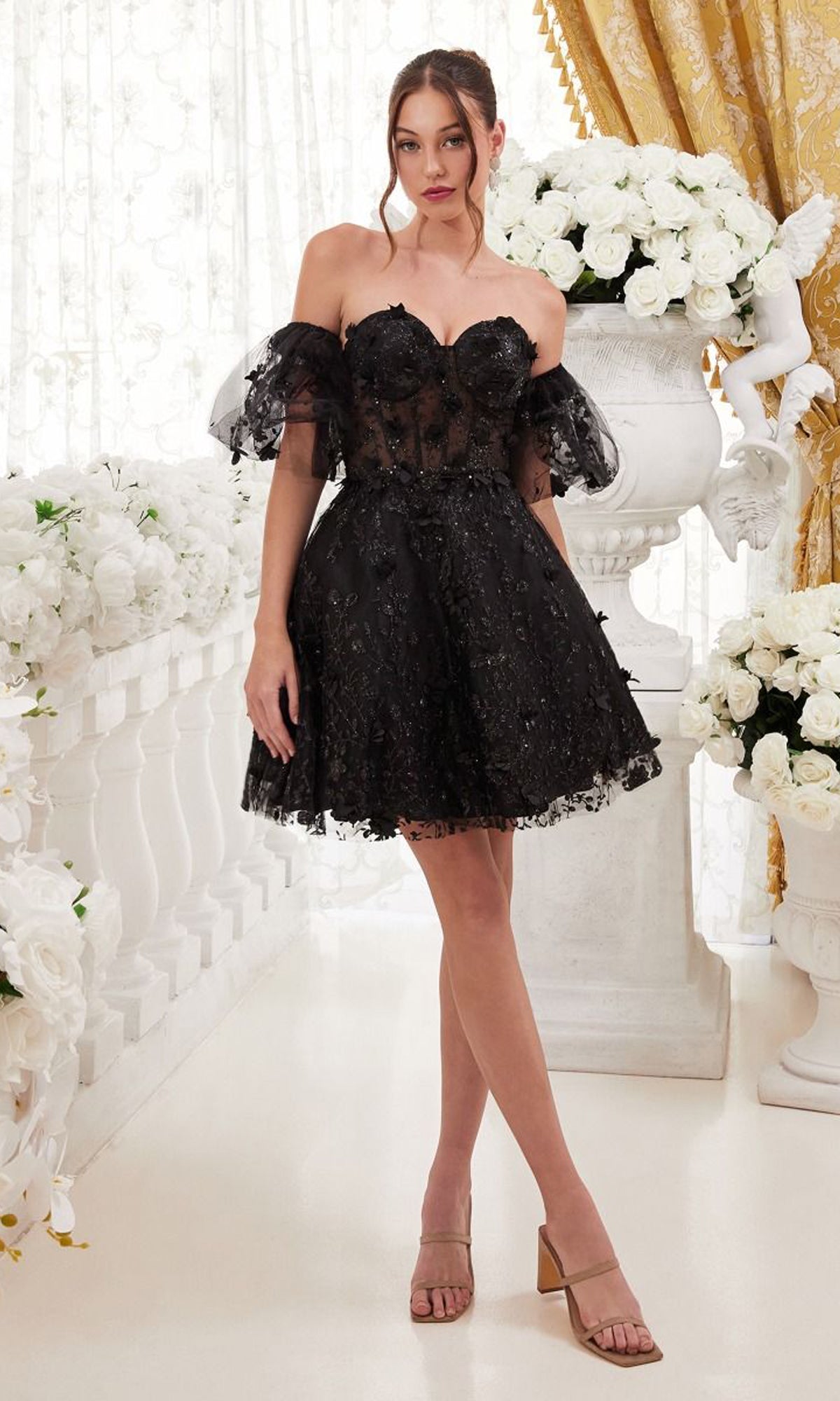 A woman stands indoors wearing the Puff-Sleeve Short Strapless Homecoming Dress KV1089 paired with nude heels. White flowers and gold curtains accent the background.